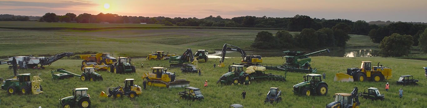 Oameni plimbându-se printre câteva tipuri de echipamente John Deere pe un câmp mare.