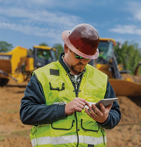 Lucrător în construcţii purtând o vestă de siguranţă verde, uitându-se la dispozitivul său mobil în faţa unui încărcător şi a unui autocamion cu benă basculantă