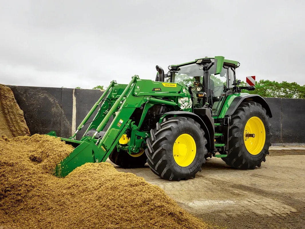 Tractor John Deere care împinge o movilă de furaje folosind cupa încărcătorului frontal, la o fermă.