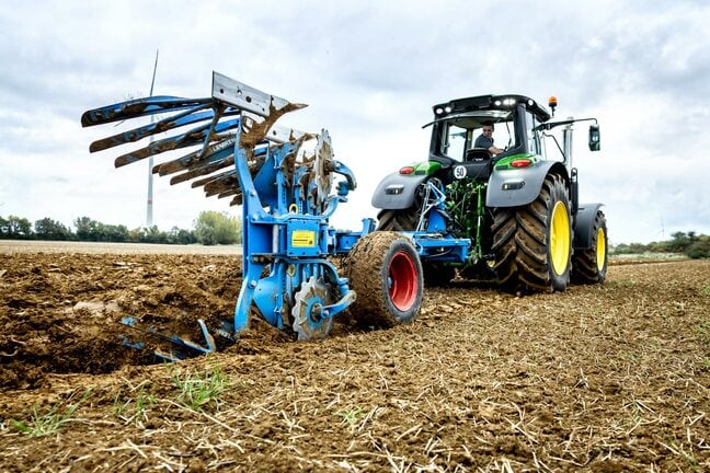 Un tractor John Deere ară cu un plug albastru reversibil pe un câmp recoltat