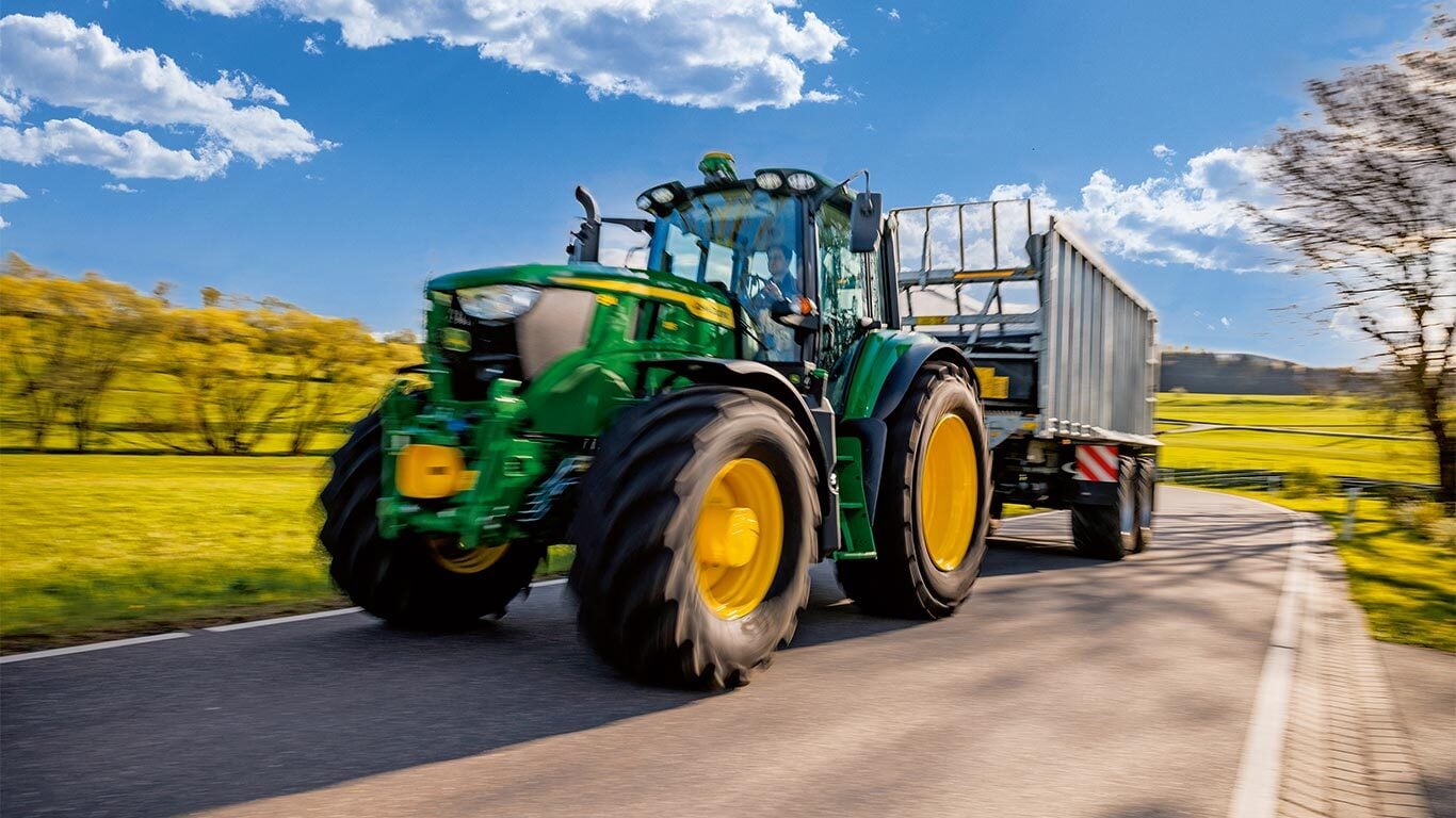 Un tractor John Deere tractează o remorcă agricolă pe un drum rural, traversând o zonă de terenuri agricole verzi, sub un cer albastru cu nori răzleţi.