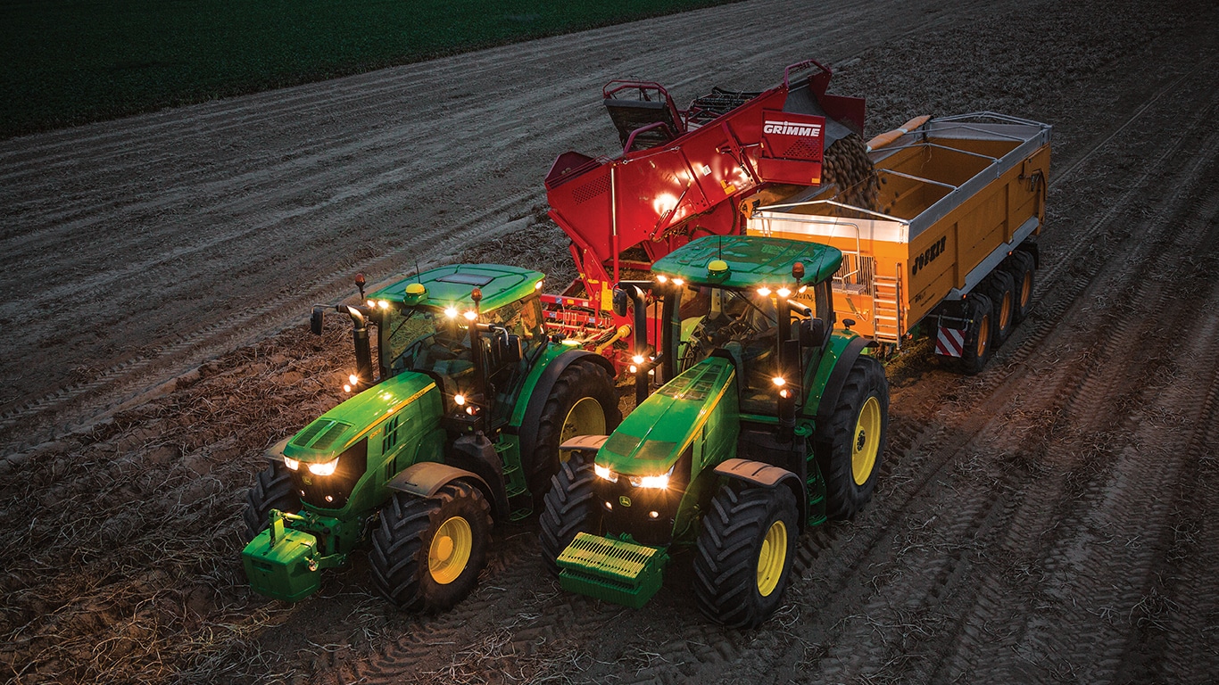Tractorul care trage o maşină de recoltat cartofi în timpul nopţii îşi descarcă cartofii într-o remorcă trasă de alt tractor