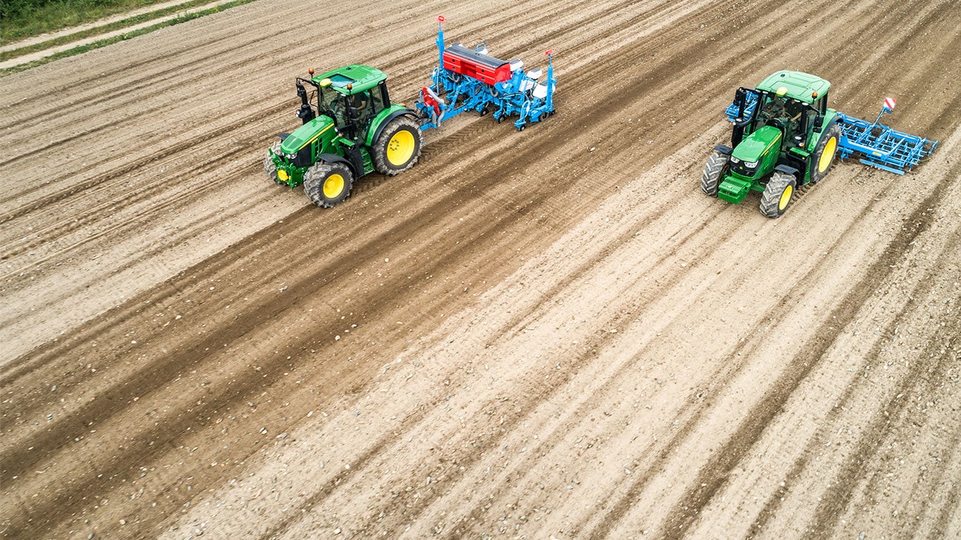 Două tractoare John Deere 6M pe un câmp la pământul gol. Unul tractează un plantator Monsomen.  Celălalt tractează o combină de pat de seminţe Lemken