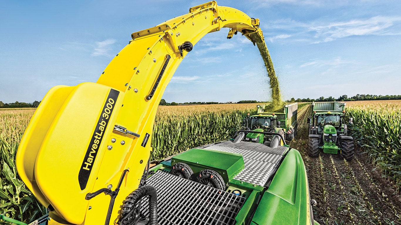 Vedere din spate a combinei autopropulsate pentru furaje John Deere Seria 9000 cu două remorci de însilozare trase de tractoare John Deere direct în spate