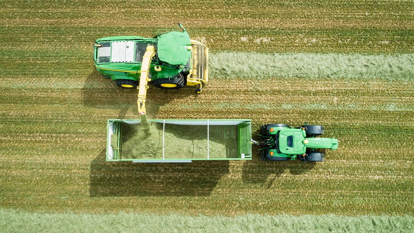 Vedere de sus a combinei autopropulsate pentru furaje John Deere Seria 8000 cu un picup de iarbă, încărcând material de siloz într-o remorcă trasă de un tractor John Deere