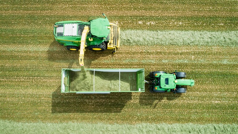 Vedere de sus a combinei autopropulsate pentru furaje John Deere Seria 8000 cu un picup de iarbă, încărcând material de siloz într-o remorcă trasă de un tractor John Deere