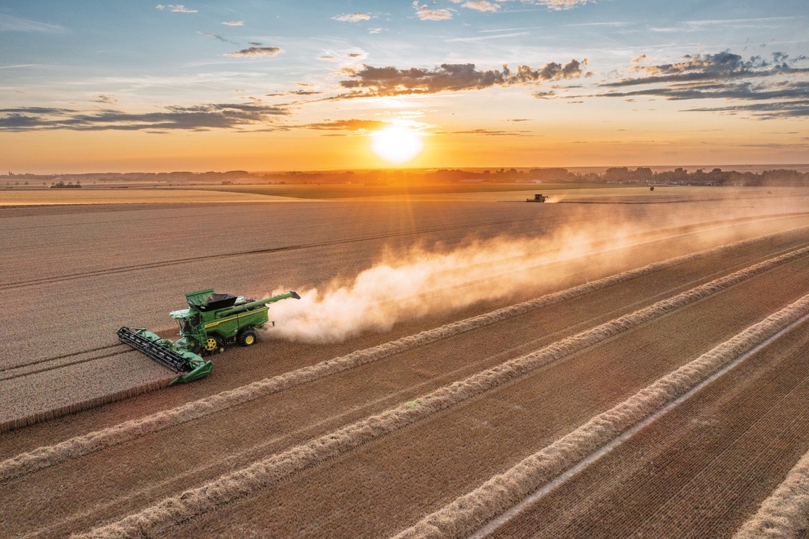 O combină recoltează grâu la apus, lăsând în urmă o dâră lungă de praf pe un teren agricol vast