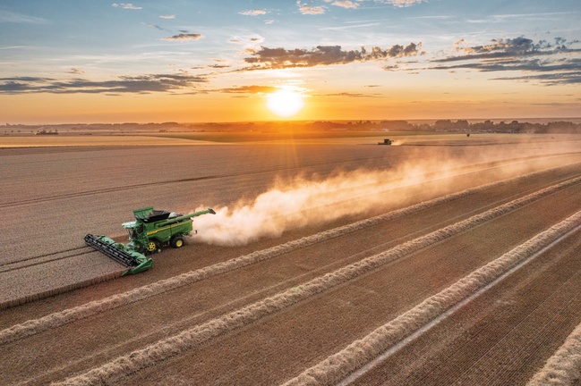 O combină recoltează grâu la apus, lăsând în urmă o dâră lungă de praf pe un teren agricol vast