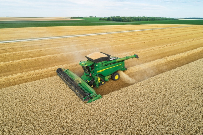 Imagine aeriană a unei combine care recoltează grâu pe câmpuri cu benzi distincte, ilustrând agricultura la scară mare