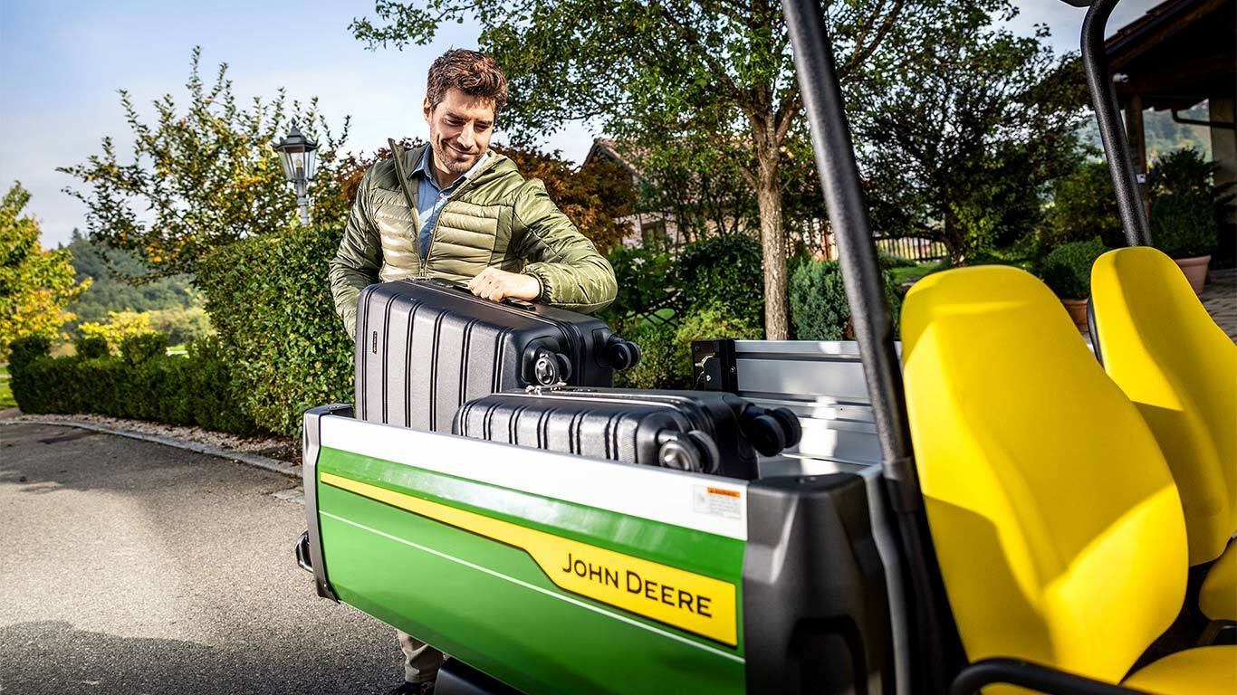 O persoană care aşază o valiză neagră mare în compartimentul de marfă al unui John Deere Gator GS cu scaune galbene, înconjurat de vegetaţie şi copaci.