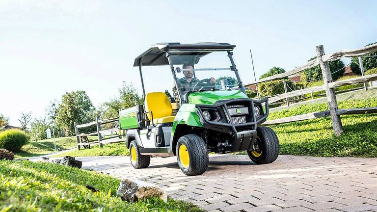 Vehicul utilitar John Deere Gator GS verde circulând pe o alee pavată printr-o zonă amenajată cu iarbă şi garduri de lemn.