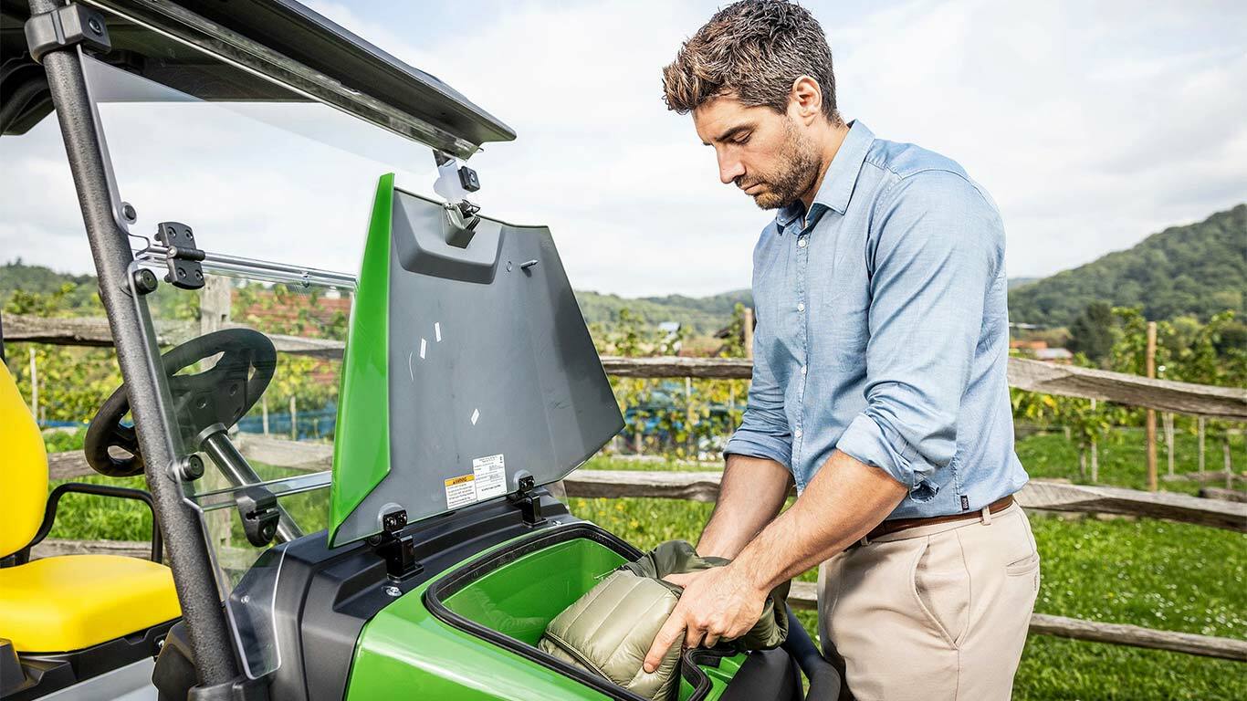 O persoană pune o geantă căptuşită în compartimentul frontal de depozitare al unui John Deere Gator GS verde, parcat afară, lângă un gard de lemn şi o viţă-de-vie.