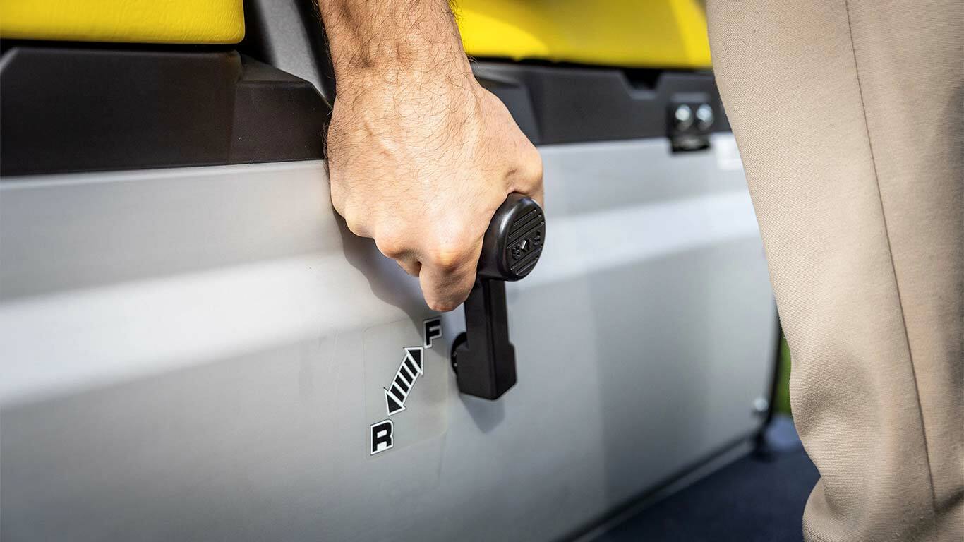 Prim-plan cu o mână care acţionează maneta selectorului de viteze al unui John Deere Gator GS, cu poziţiile de înaintare şi marşarier lângă scaunele galbene.
