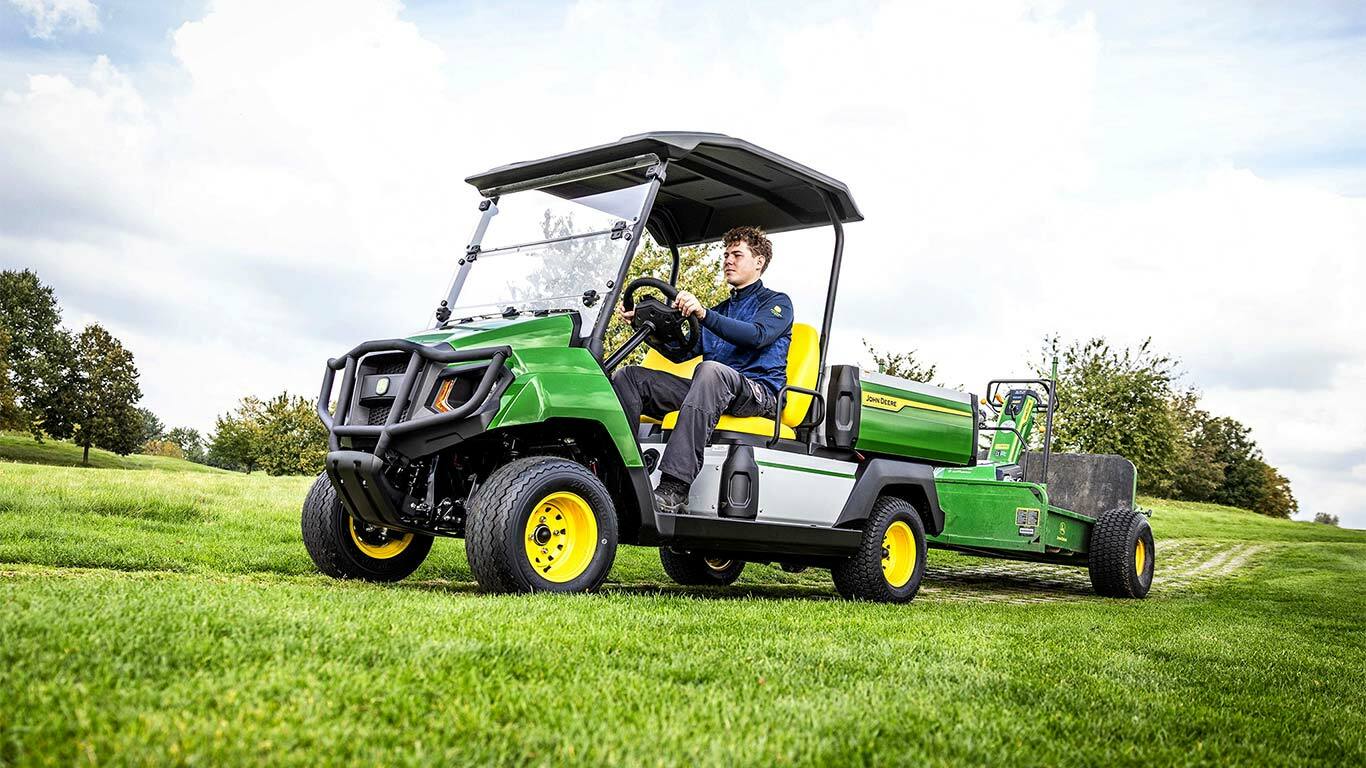 Vehicul utilitar electric John Deere Gator verde tractează echipamente pe un câmp cu iarbă, sub un cer parţial înnorat.