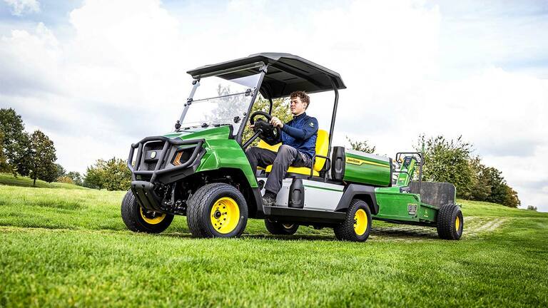 Vehicul utilitar electric John Deere Gator verde tractează echipamente pe un câmp cu iarbă, sub un cer parţial înnorat.