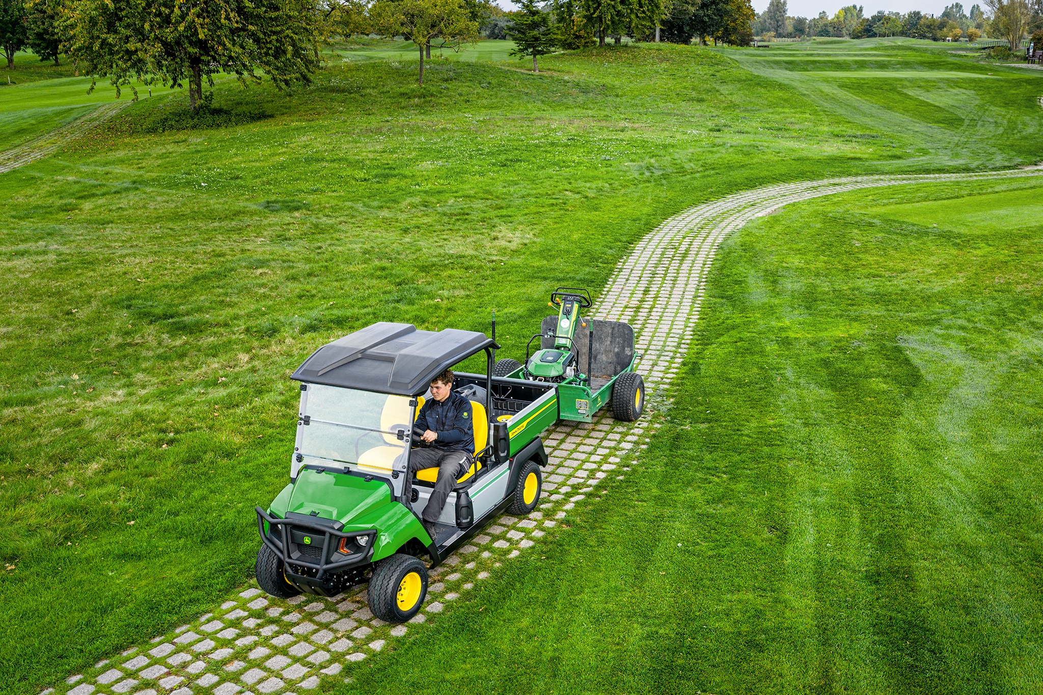 John Deere Gator GS tractând echipamente pe o alee cu iarbă dintr-un teren de golf John Deere Gator GS tractând echipamente pe o alee cu iarbă dintr-un teren de golf