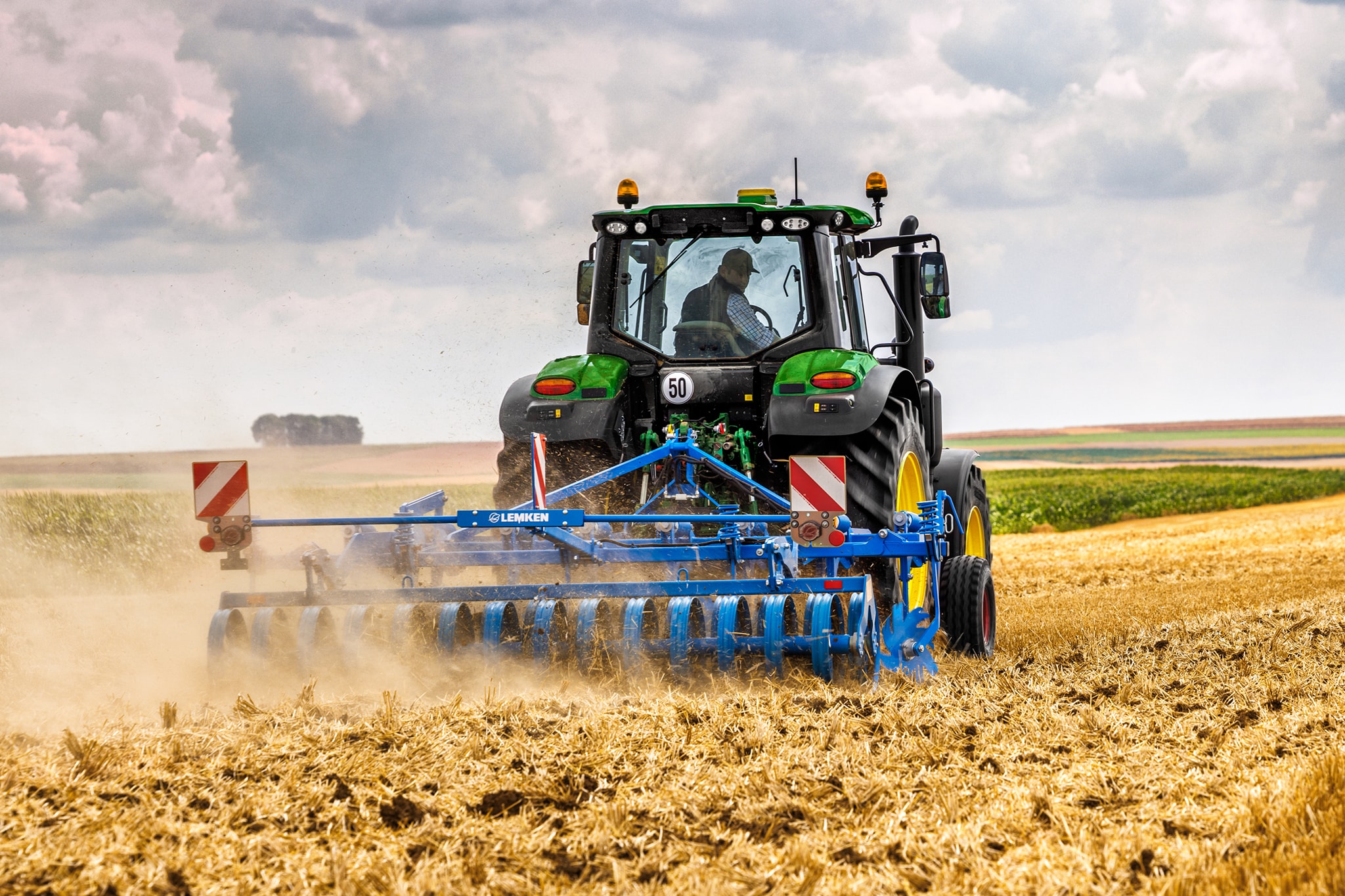 Tractorul John Deere seria 6M care cultivă un câmp recoltat cu o grapă cu discuri, vedere din spate în condiţii de câmp cu praf.