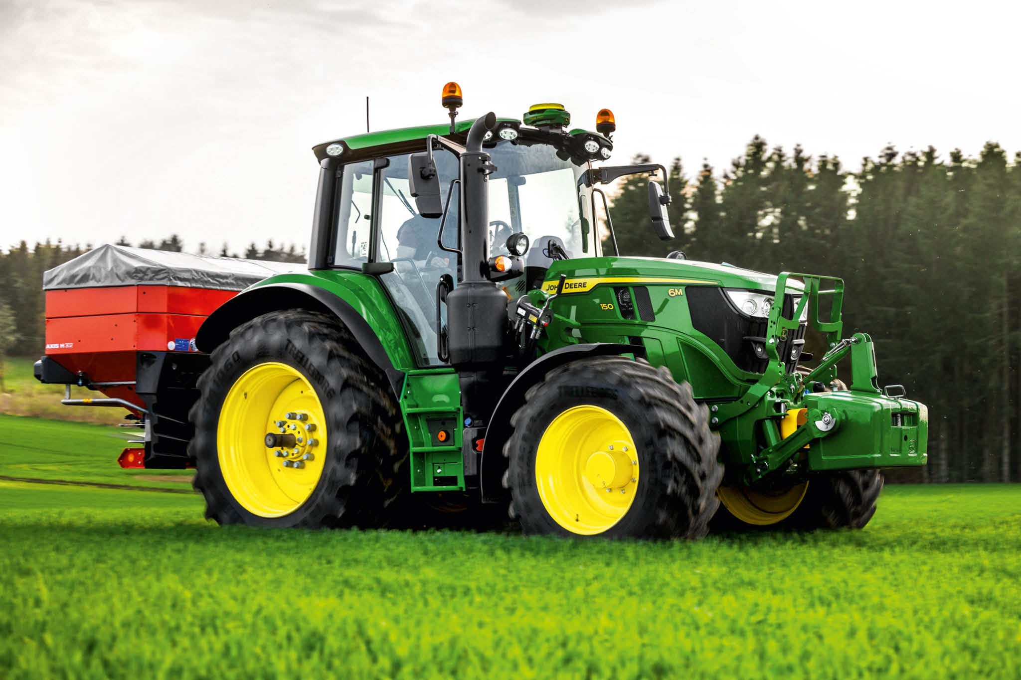 Tractor John Deere Seria 6M cu împrăştietor montat în spate care lucrează pe iarbă la marginea unei păduri.