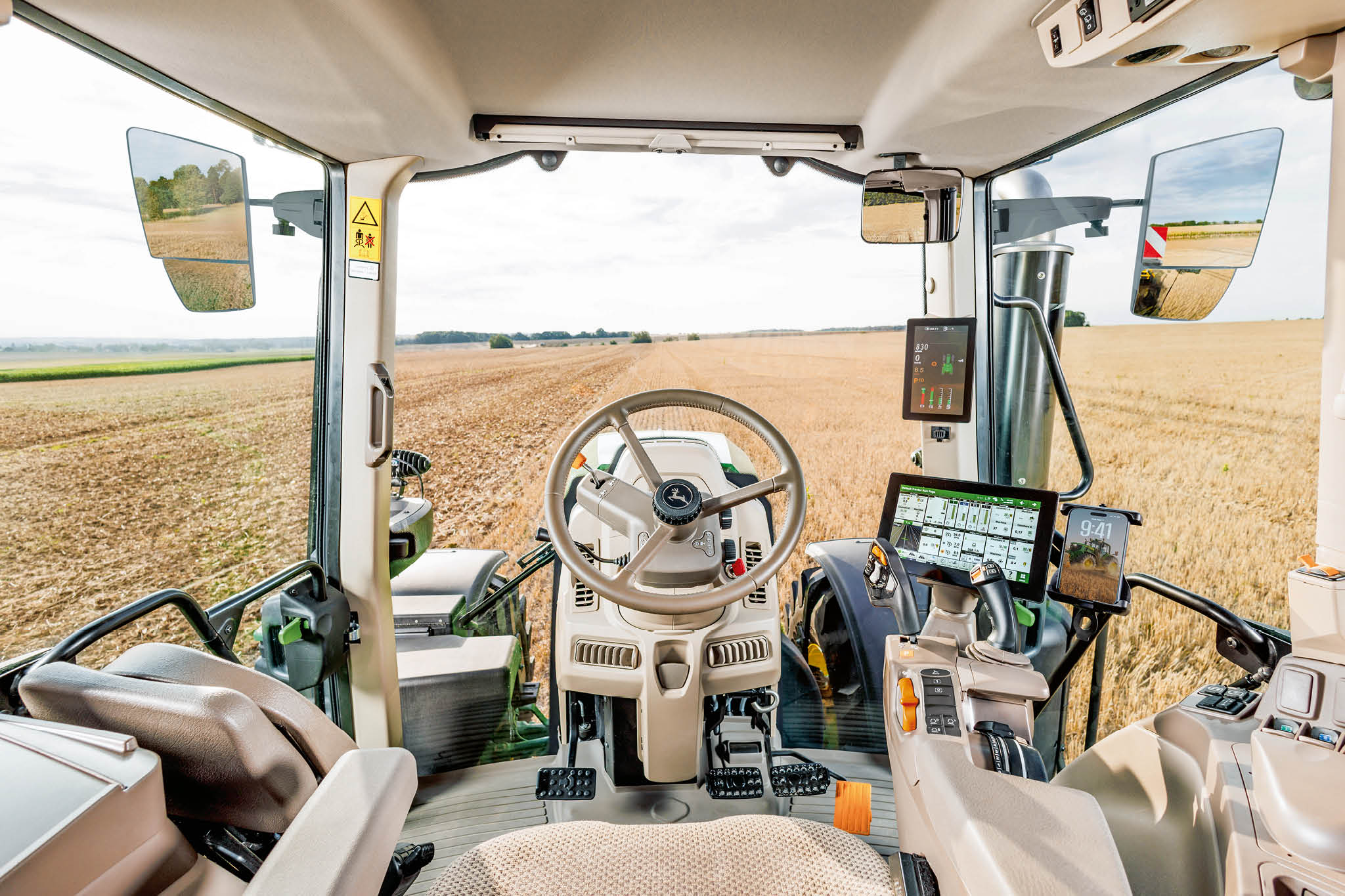 Interiorul cabinei tractorului John Deere cu volan, afişaje de control digital şi vizibilitate spre câmp prin parbriz în timpul lucrărilor agricole.