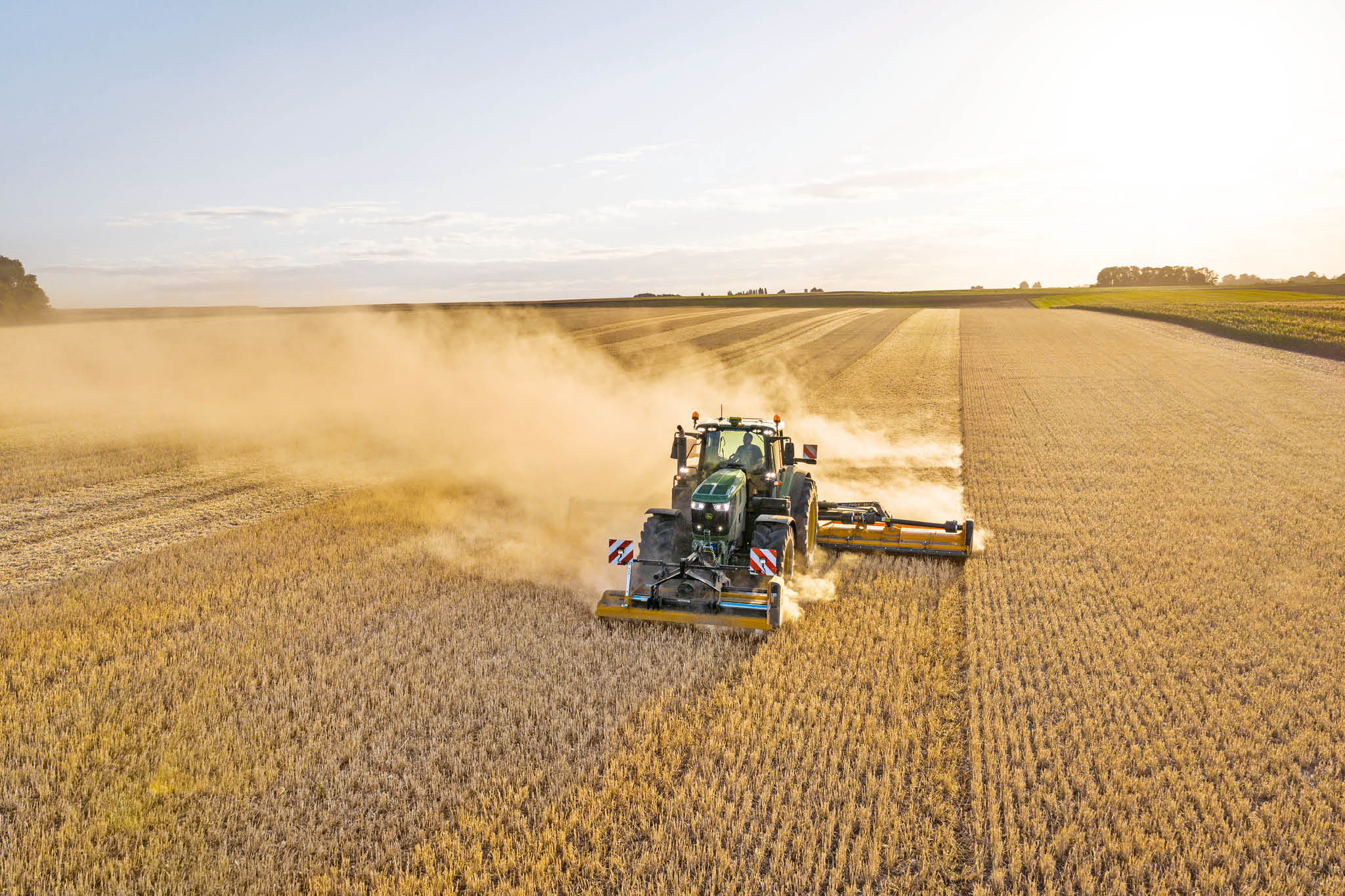 Un tractor John Deere seria 6M tractează o cisternă de decantare de-a lungul unui drum rural mărginit de iarbă şi câmpuri.
