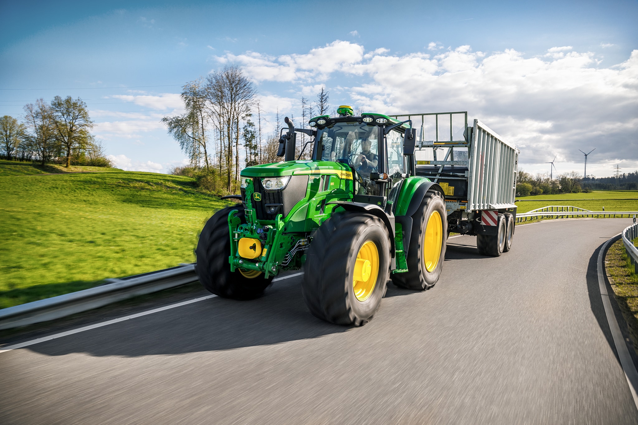 Un tractor John Deere seria 6M transportă o cu obloane înalte pe un drum rural asfaltat.