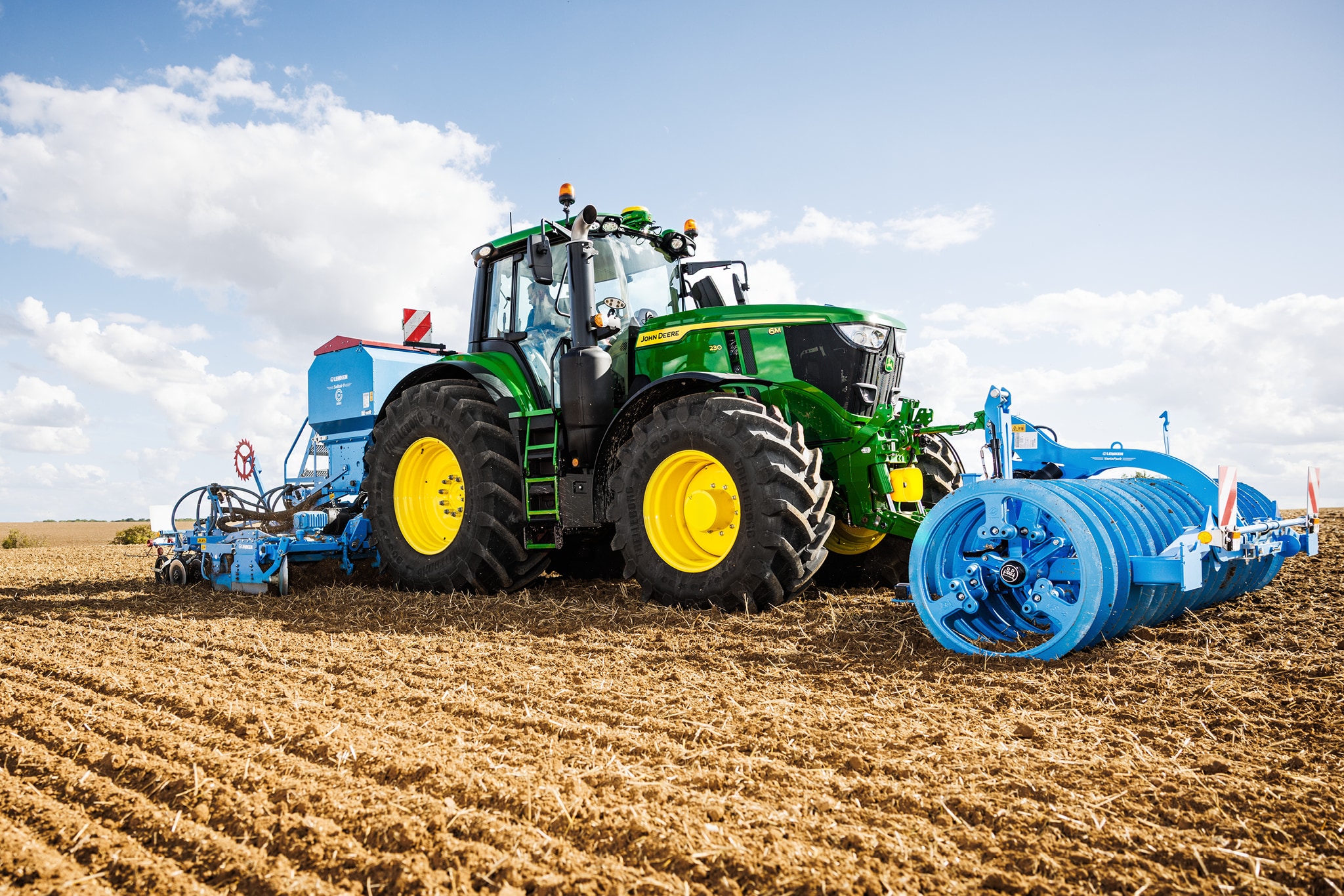 Un tractor John Deere seria 6M cultivă câmpul cu un instrument cu role albastre de mari dimensiuni ataşat.