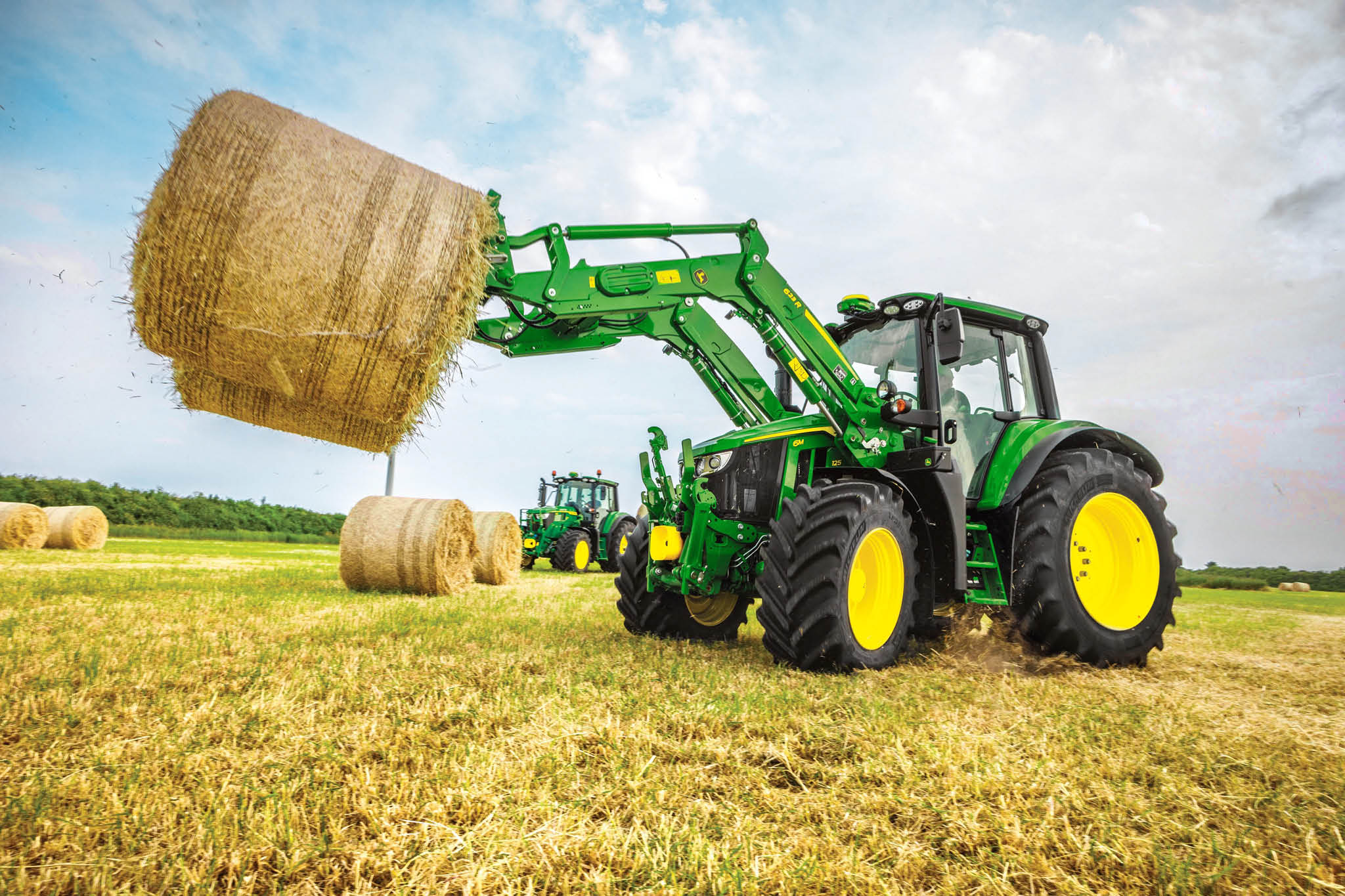 Tractorul John Deere seria 6M ridică un balot rotund de fân de dimensiuni mari cu un încărcător frontal într-un câmp deschis.