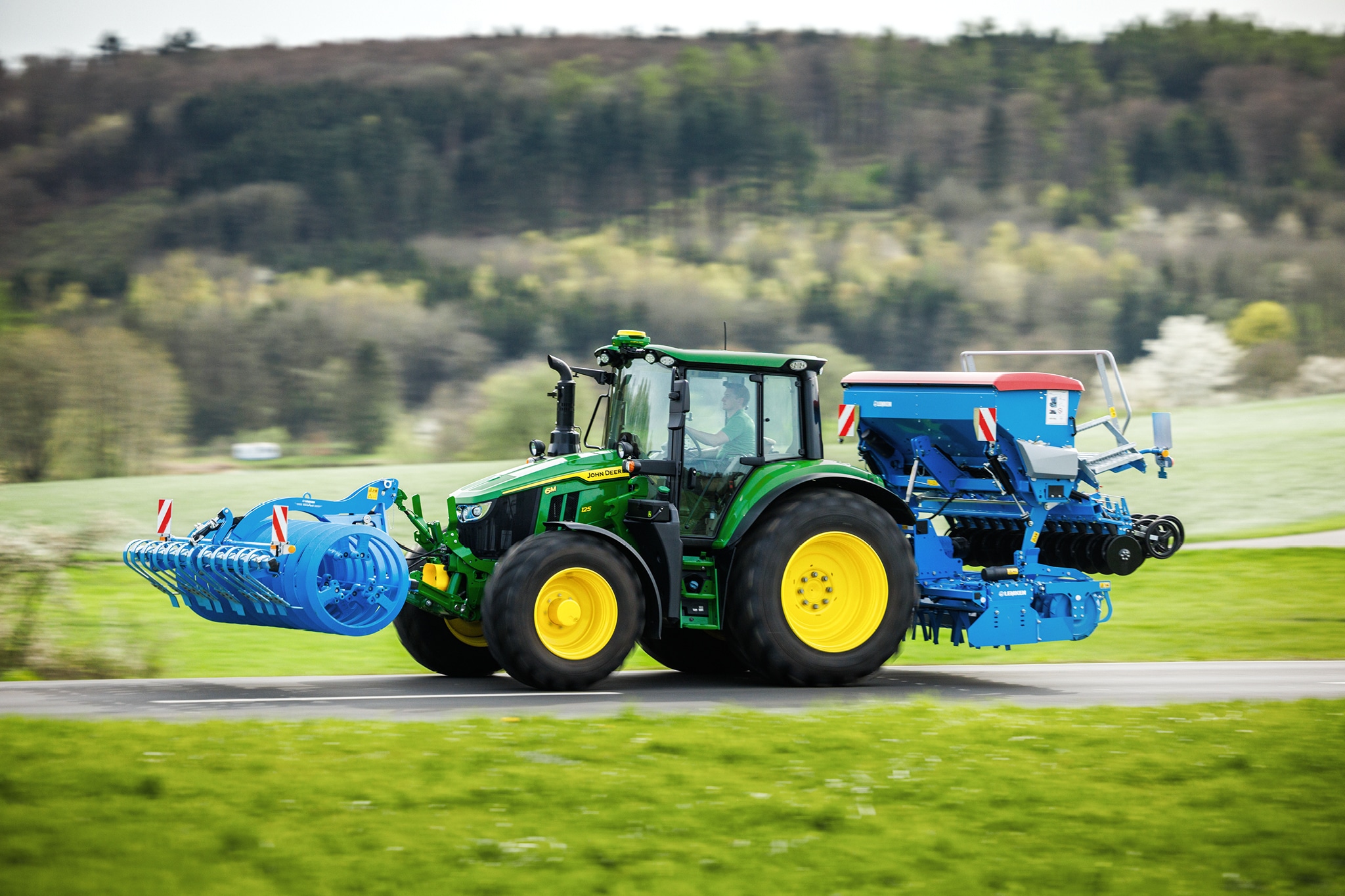 Un tractor John Deere seria 6M transportă un instrument montat frontal şi o semănătoare posterioară pe un drum rural.