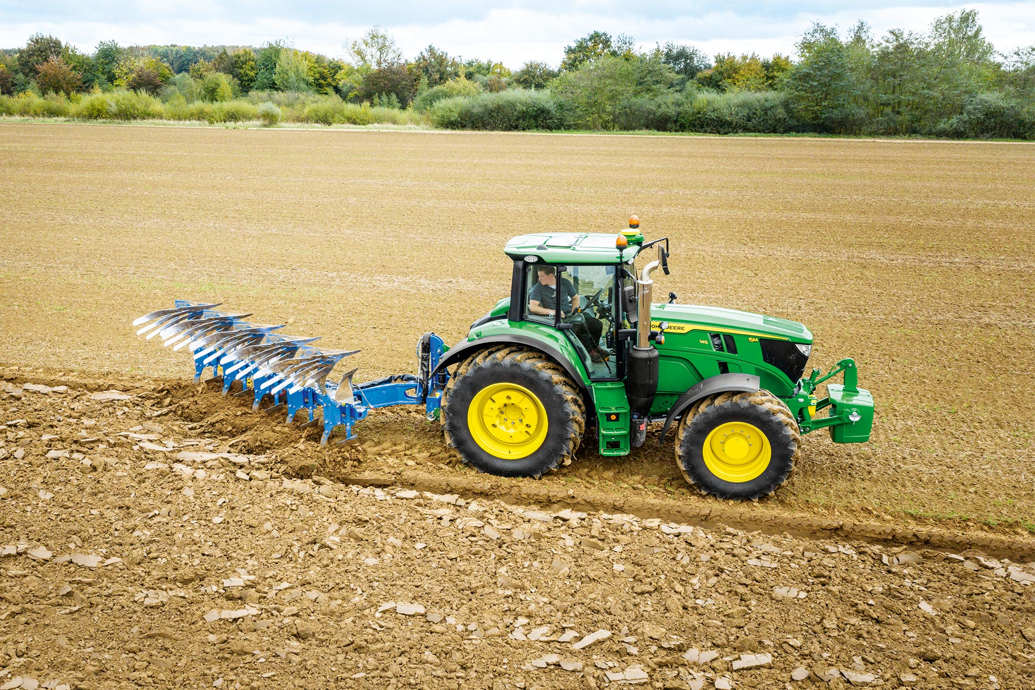 Tractor John Deere seria 6M care lucrează pe un câmp arat cu un instrument de arat montat în spate, sub cerul înnorat.