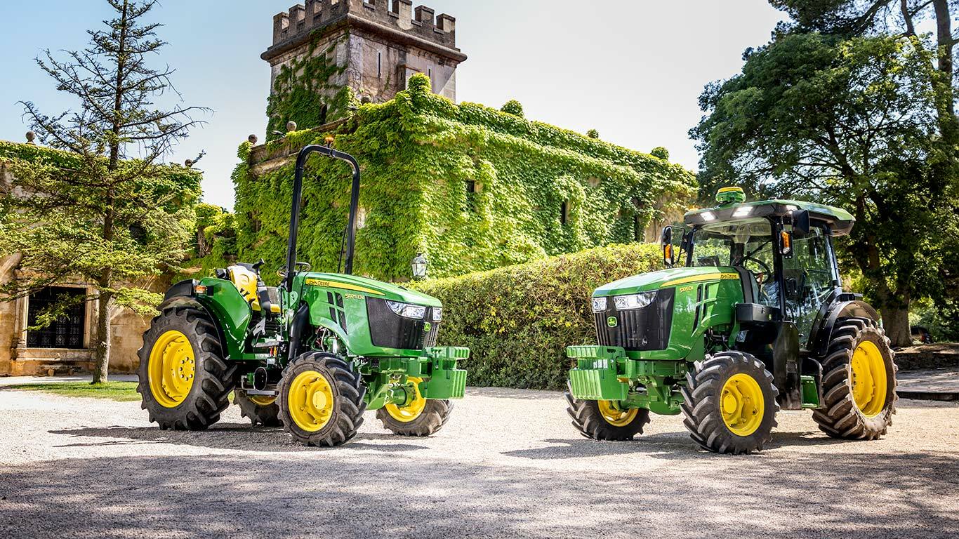 Tractorul John Deere 5075EN, în verde şi galben, trece pe un drum de pământ dintr-o livadă, scăldat în lumina soarelui şi înconjurat de pomi fructiferi.