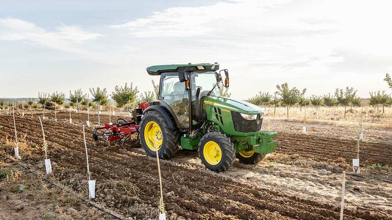 Tractor John Deere 5075EN cultivă solul pe un câmp deschis cu cer senin și rânduri plantate.