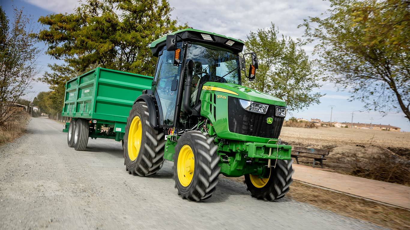 Tractor John Deere 5075EN tractează o remorcă verde pe un drum rural cu copaci în fundal.