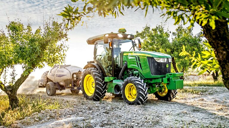 Tractorul John Deere 5090EN, în verde şi galbe, trage o autocisternă de stropit prin livadă într-o zi însorită.