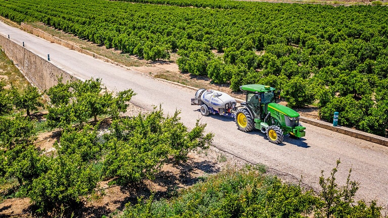 Tractorul John Deere 5090EN, în verde şi galben, tractează o autocisternă de stropit pe un drum pavat între rândurile mari ale unei livezi, sub un cer senin.