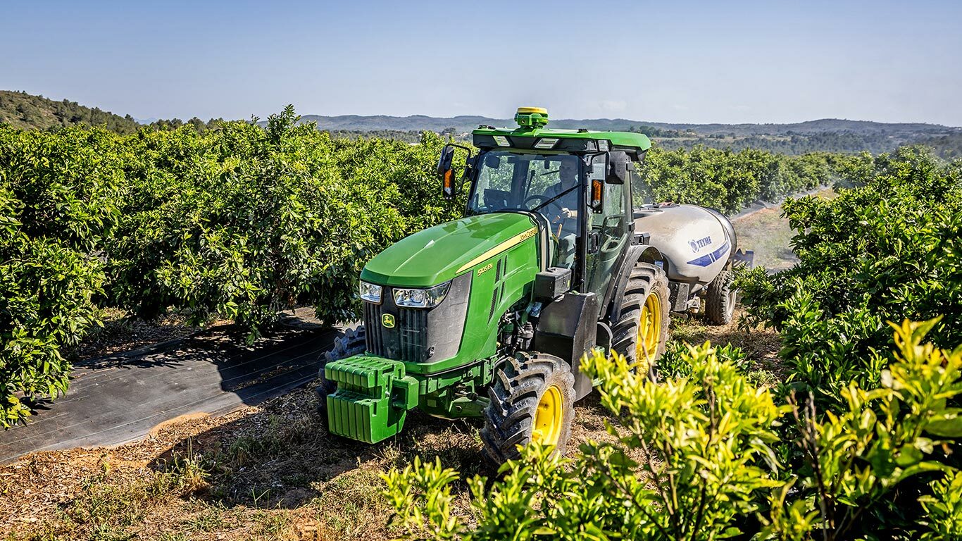Tractorul John Deere 5105EN, în verde şi galben, tractează o autocisternă de stropit într-o livadă bogată, cu dealuri domoale în fundal.