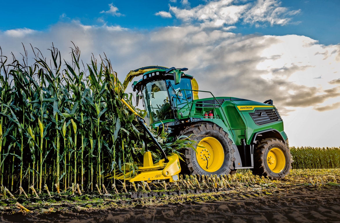 Combină de furaje John Deere care recoltează porumb pe câmp sub un cer albastru cu nori.