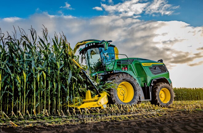 Combină de furaje John Deere care recoltează porumb pe câmp sub un cer albastru cu nori.