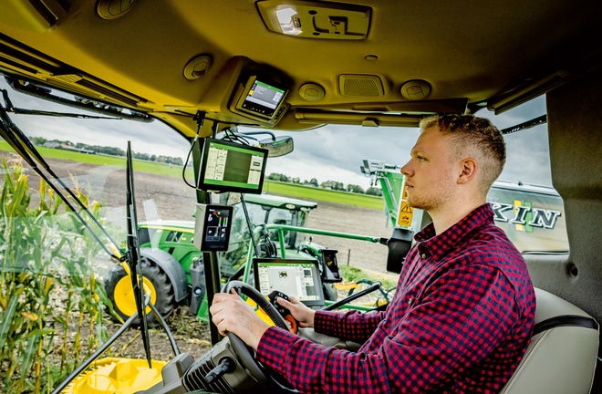 Fermier care utilizează utilaje agricole avansate cu mai multe ecrane digitale în cabina unei combine de furaje autopropulsate, cu tehnologie agricolă modernă pe un câmp.