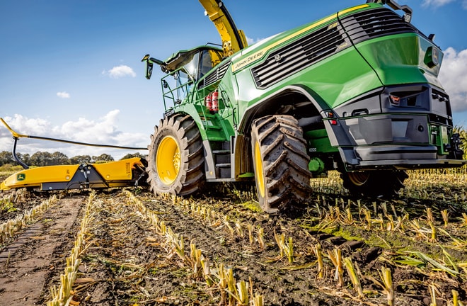 Combină de furaje John Deere care lucrează într-un lan de porumb recoltat, cu pneuri mari ale utilajului şi mecanism de tăiere pe un cer albastru senin.